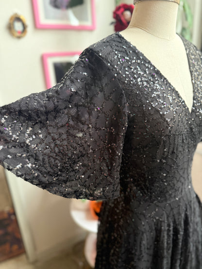 A black spiderweb sequined dress displayed on a mannequin torso in an indoor setting. The dress features a V-neckline and voluminous, wide flared sleeves with dense sequins throughout.