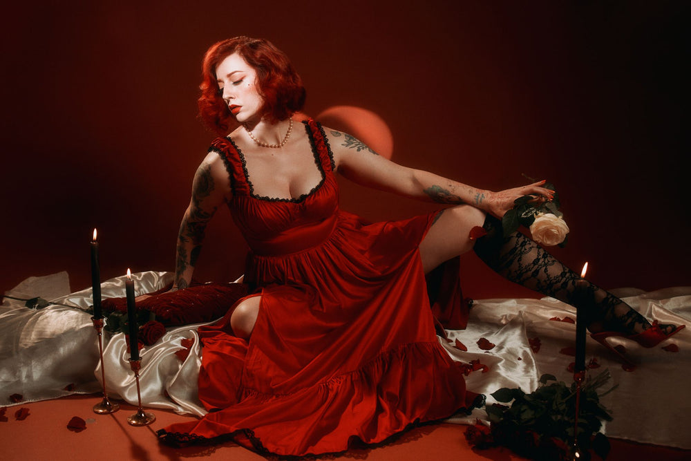 Model showcasing the Cosette Satin dress in crimson red with lace trim and thigh high slits, surrounded by rose petals and candles.