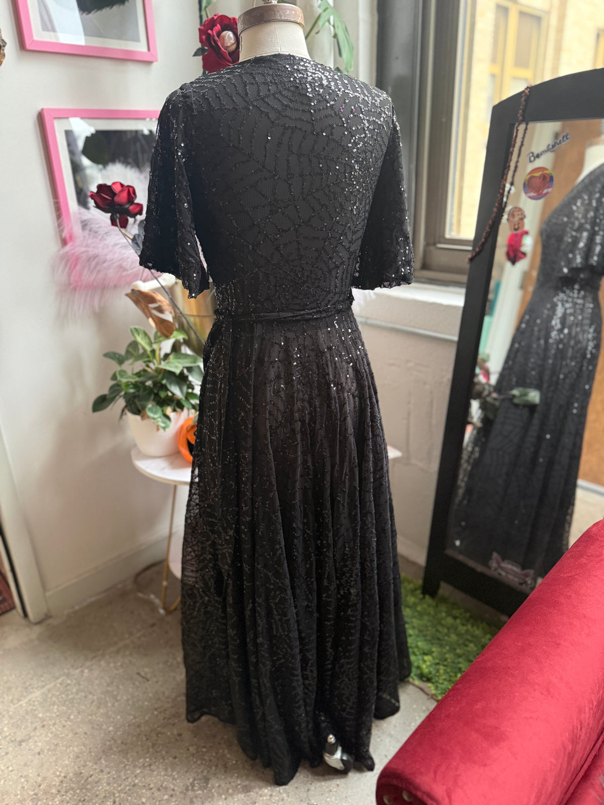A black sequined evening gown displayed on a mannequin in an indoor room, viewed from the back. The dress has short sleeves, a fitted waist, and a full skirt with dense sequin embellishment.
