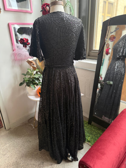 A black sequined evening gown displayed on a mannequin in an indoor room, viewed from the back. The dress has short sleeves, a fitted waist, and a full skirt with dense sequin embellishment.