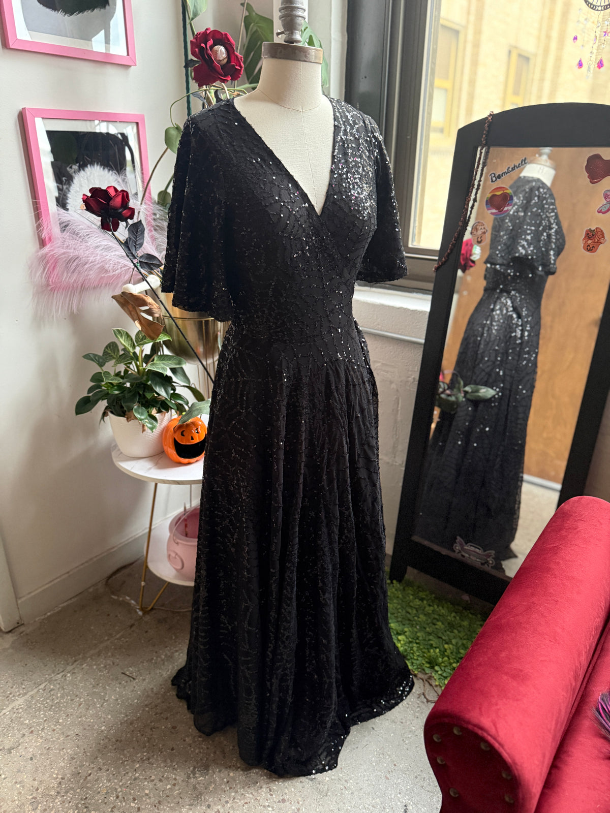 A floor-length black spiderweb sequined gown displayed on a mannequin in an indoor room. The dress has short sleeves, a deep V-neckline, and a flared skirt.