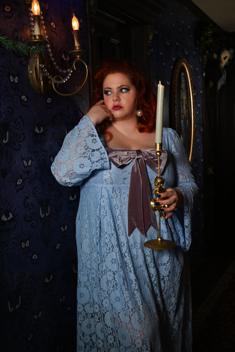 Model in Bippity Boppity Blue lace maxi dress with bell sleeves and removable mauve bow, holding a candle in a vintage setting.