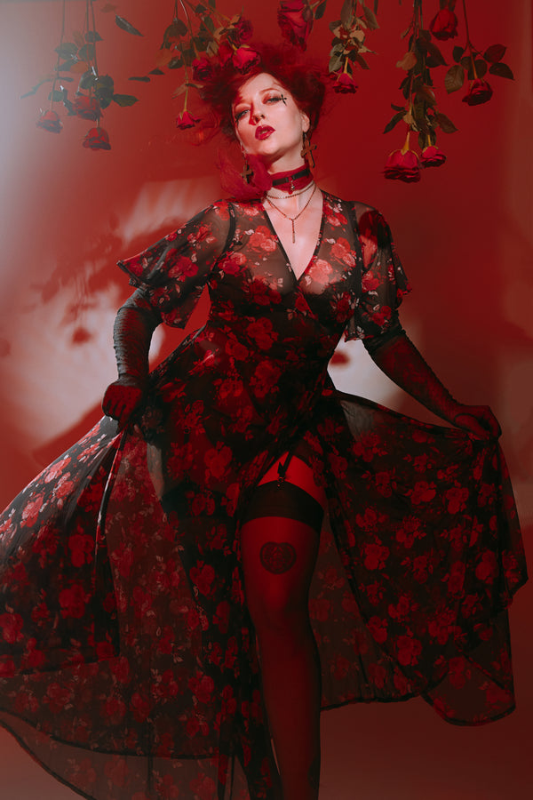 Model in dramatic rose cluster mesh wrap dress, showcasing pockets and romantic style against a red floral background.