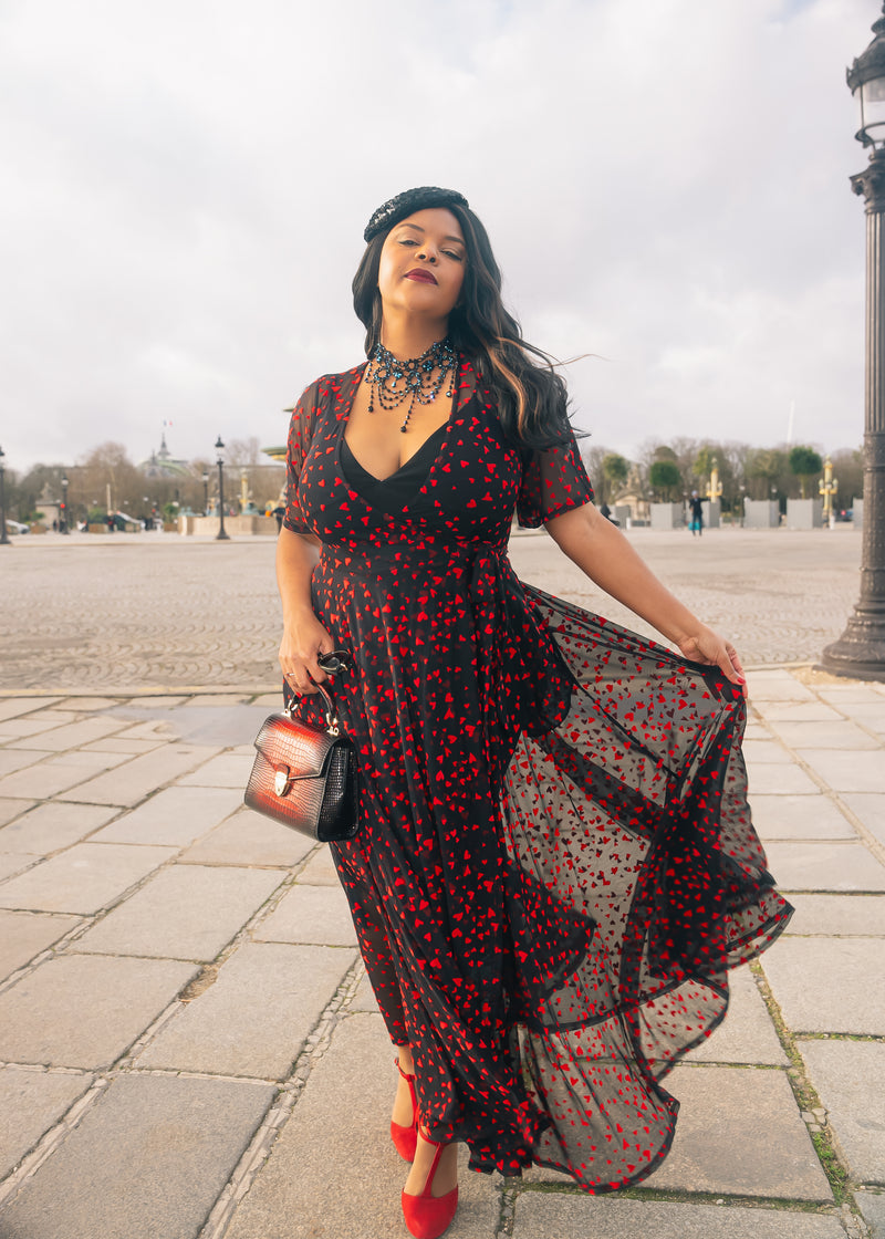 Woman in a black mesh wrap maxi dress with red hearts, twirling in a city square, perfect for Valentine's Day.