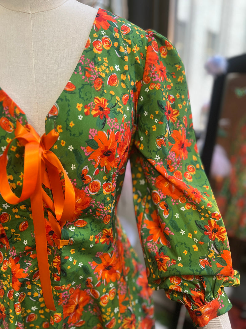 60s inspired mini floral dress with bishop sleeves in vibrant green and orange floral print, featuring a lace up front and heart buttons.