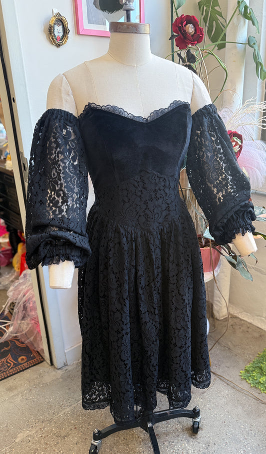 A black lace off-the-shoulder dress displayed on a cream mannequin torso in a boutique setting. The dress features a structured corset-style bodice, a flared skirt, and sheer lace puff sleeves with ruffled cuffs.