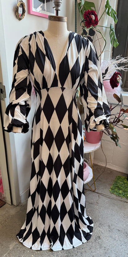 A long black and white harlequin diamond-pattern dress displayed on a mannequin in a room with a window, mirror, and plants.