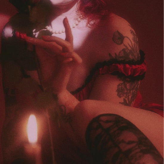 Woman in a romantic crimson dress with lace trim, surrounded by candles and holding a rose.