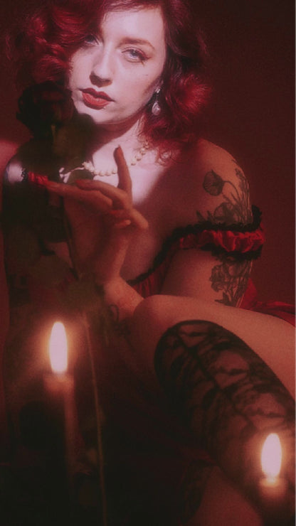 Woman in a romantic crimson dress with lace trim, surrounded by candles and holding a rose.