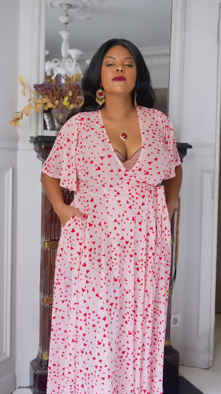 Model in pink mesh maxi wrap dress with flocked red hearts and pockets, perfect for Valentine's Day.